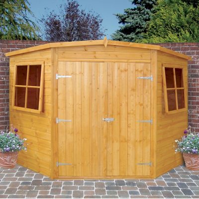 A wooden corner garden shed with a natural finish, featuring double doors, two windows, and a sloped roof, set against a brick wall and greenery.