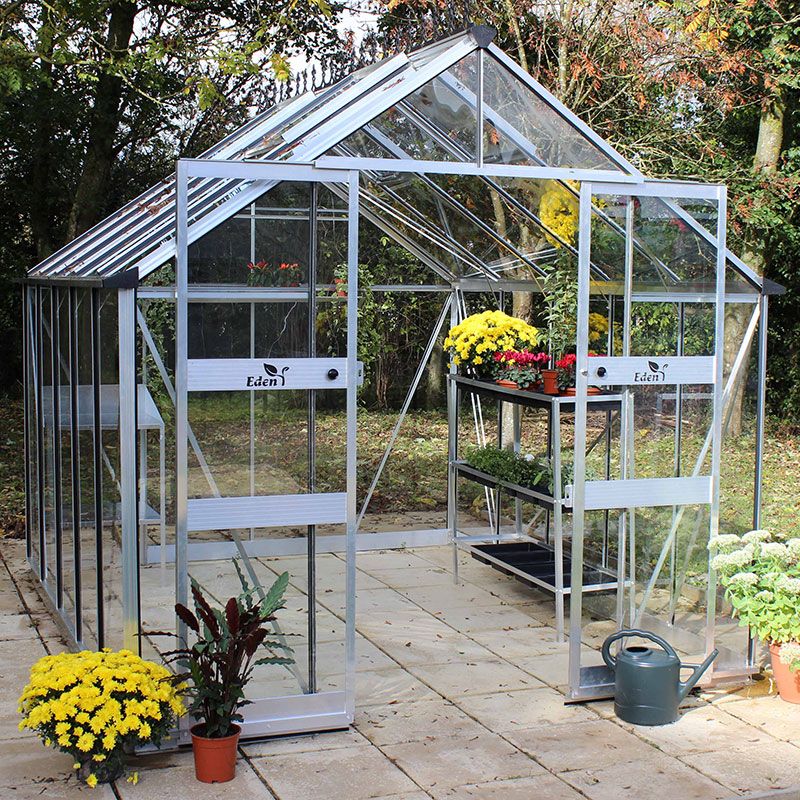 8' x 10' Halls Cotswold Blockley greenhouse featuring a silver aluminum frame, toughened glass panels, double doors, and potted yellow flowers on shelves and beside the entrance.