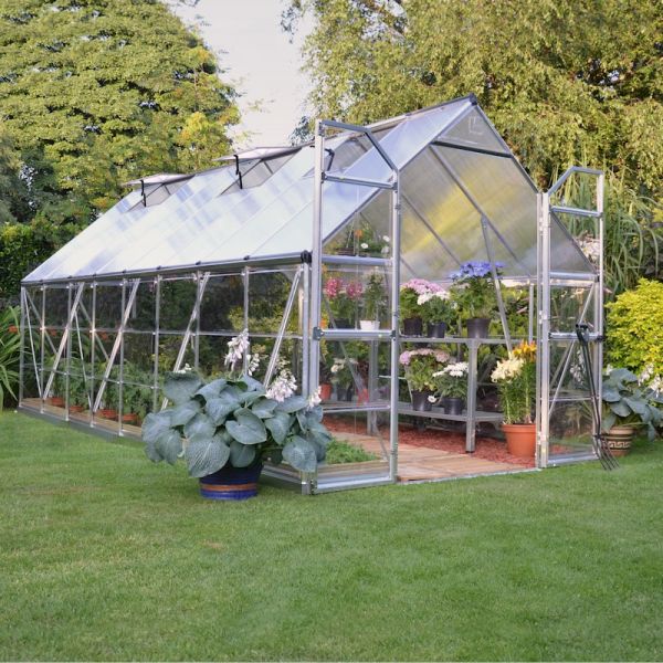 Silver aluminum greenhouse with clear polycarbonate panels, measuring 8' x 16', featuring double doors, a sloped roof, and surrounded by lush greenery and potted plants.