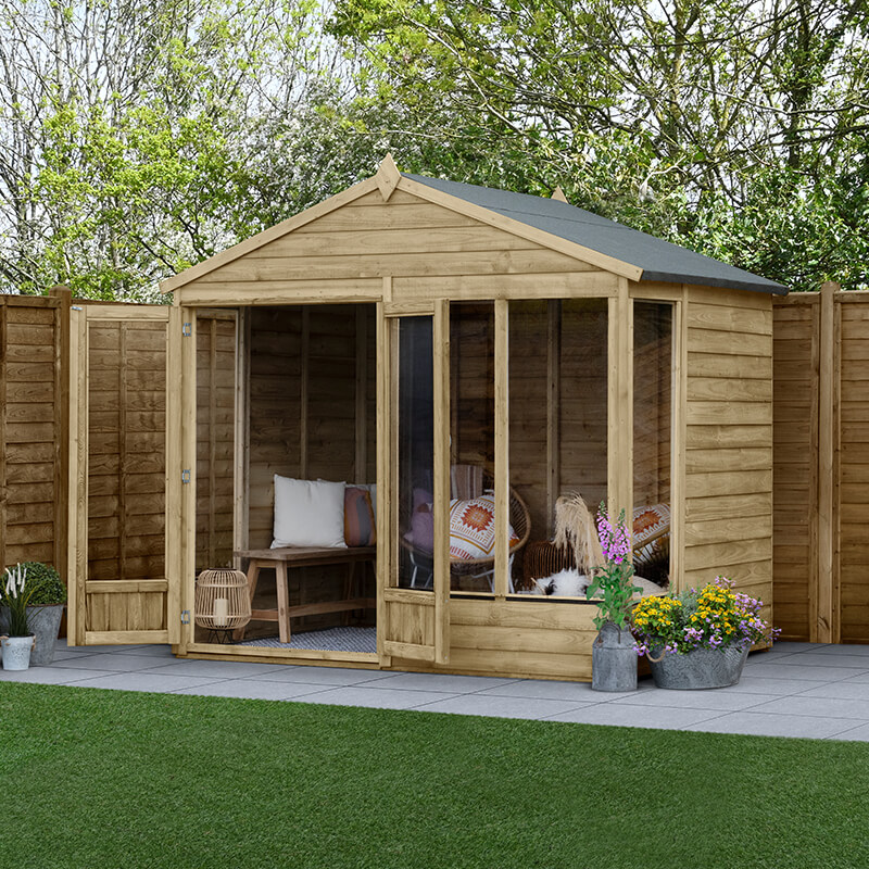 A wooden summer house with a double door and four windows, featuring a sloped apex roof, surrounded by greenery and potted flowers on a stone patio.