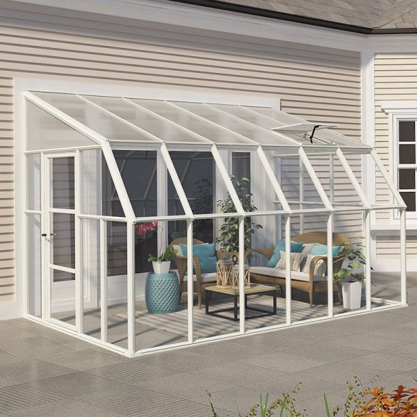 White walk-in greenhouse with a glass roof and walls, featuring a spacious interior with potted plants, a coffee table, and seating, set against a light-colored building.