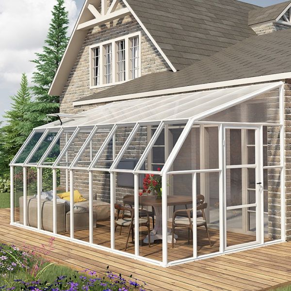 White walk-in greenhouse with a sloped roof, featuring transparent polycarbonate panels, double doors, and a wooden deck, set against a stone house and surrounded by greenery.