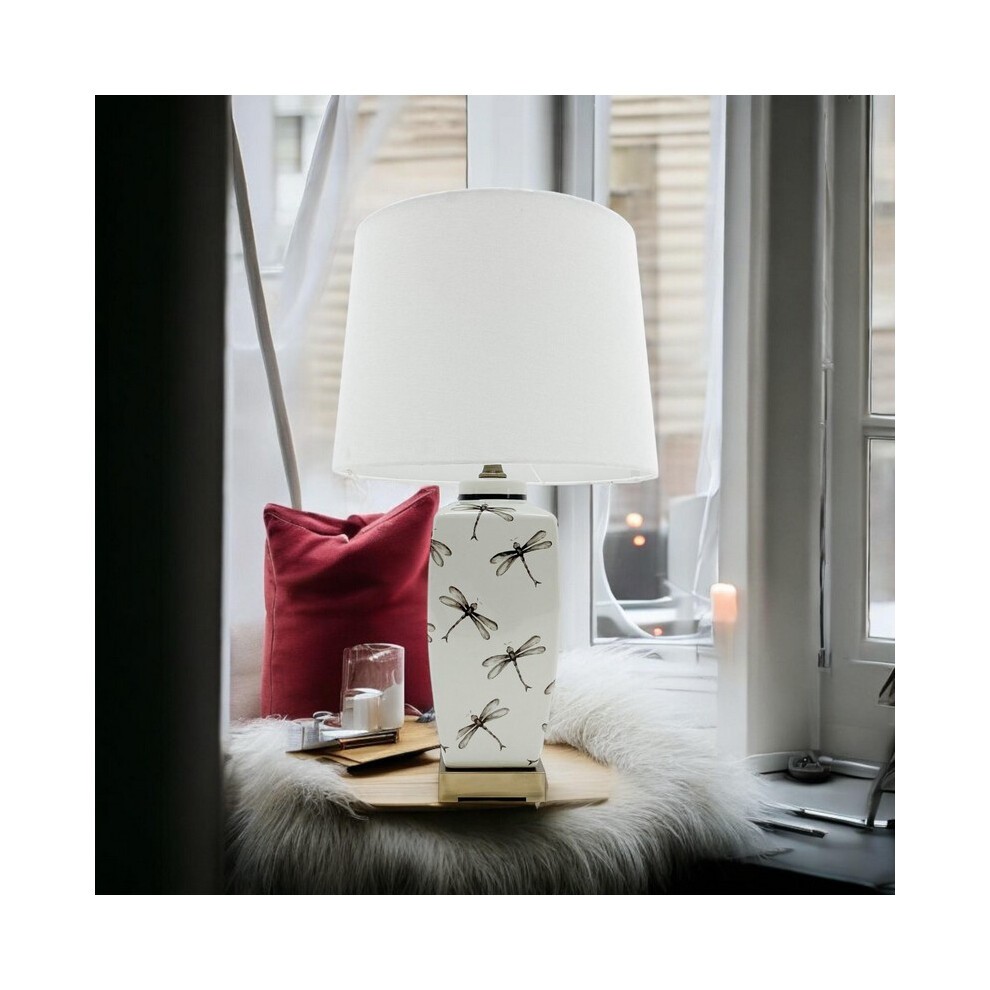Ceramic table lamp with a white base featuring black dragonfly designs, topped with a white fabric shade, set on a cozy window sill with a red cushion.