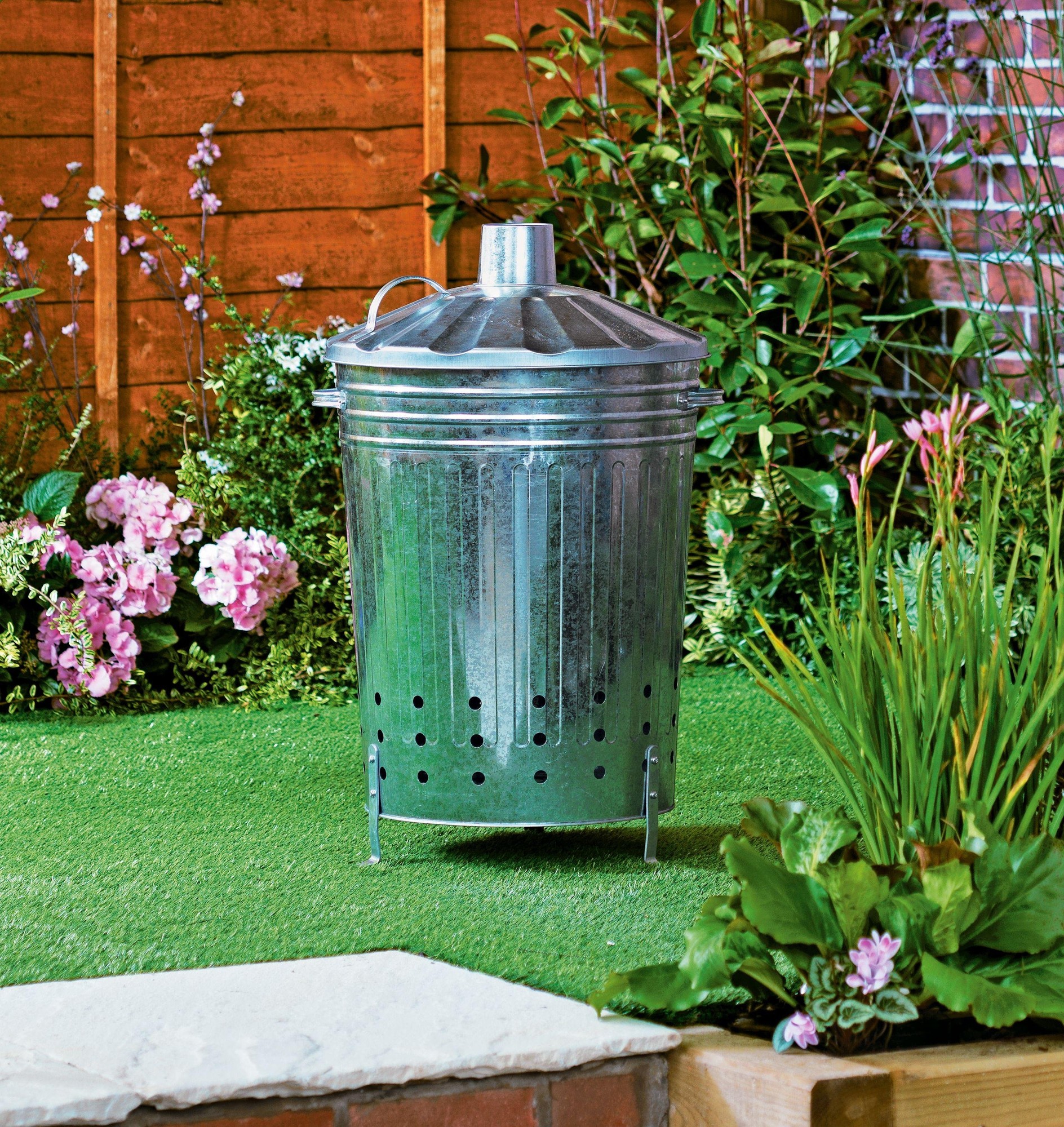 Galvanised grey garden incinerator with a domed lid, vent holes, and sturdy legs, set against a backdrop of lush greenery and colorful flowers.