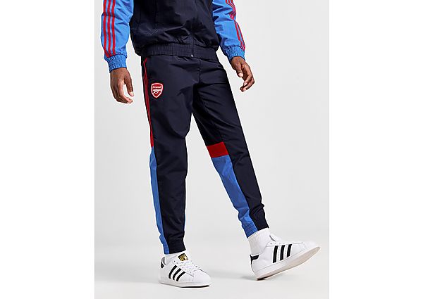 Navy woven tracksuit bottoms featuring red and blue side panels, an Arsenal logo on the thigh, and a tapered fit, paired with white sneakers.