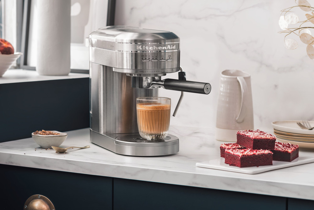 Stainless steel KitchenAid ARTISAN semi-automatic espresso machine on a marble countertop, with a glass of espresso and red velvet cake squares nearby.