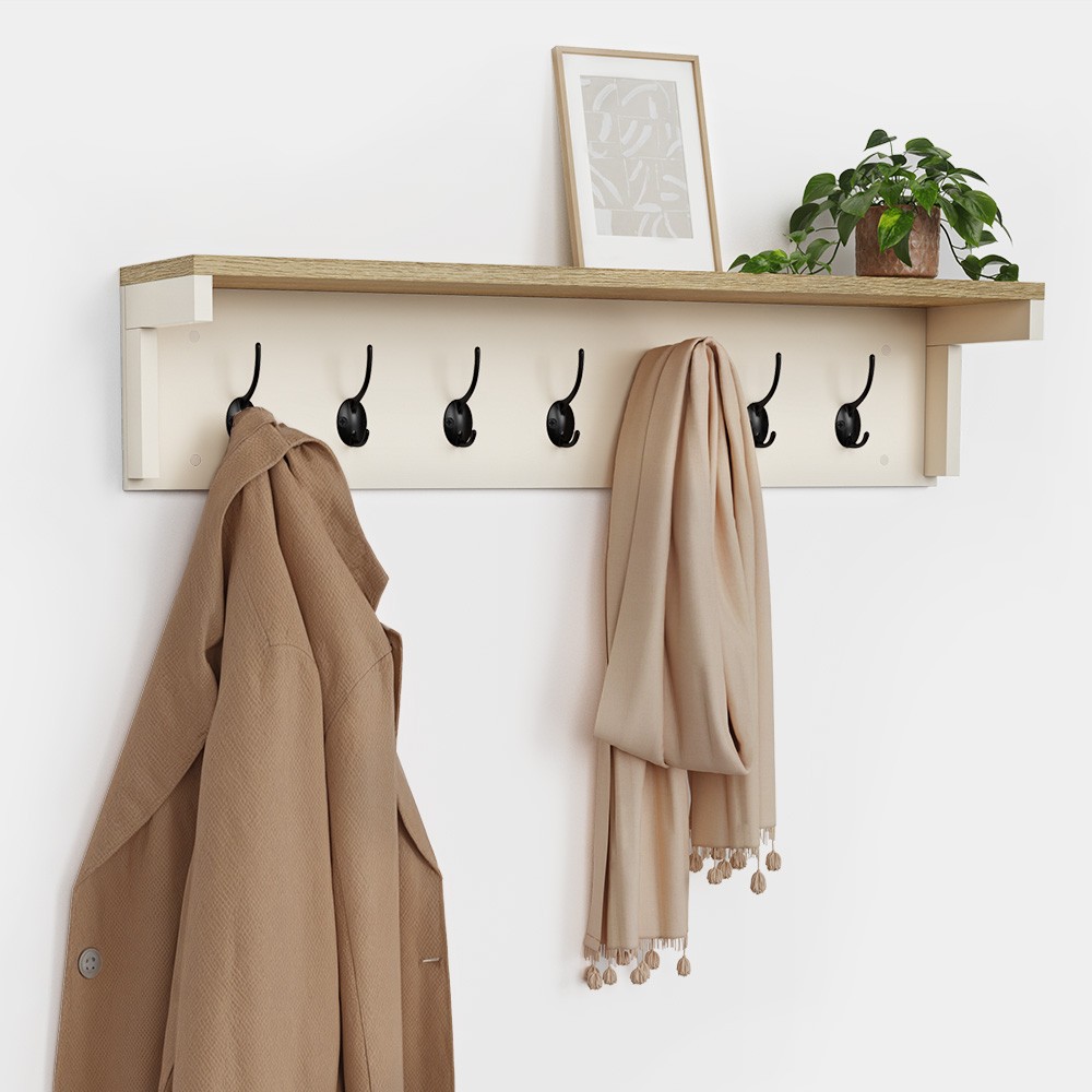 Cream wall-mounted coat rack featuring a wooden shelf, six black hooks, and a decorative plant, with a framed picture above and a beige coat and scarf hanging.