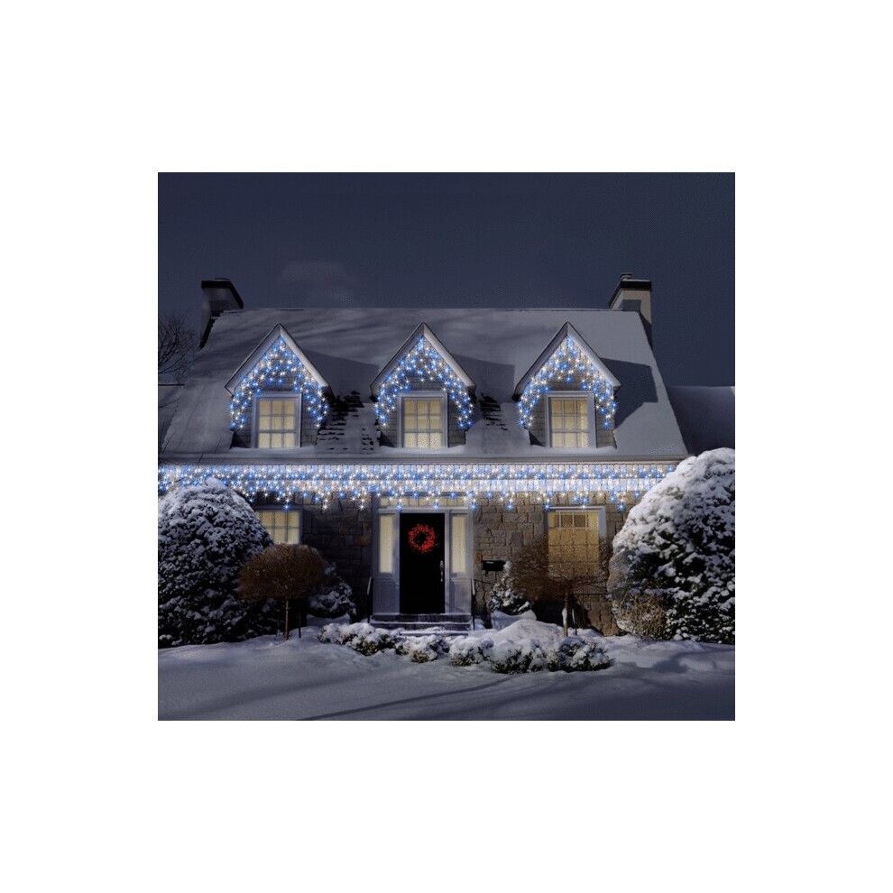 Bright white and blue LED icicle lights hang from a snow-covered roof, illuminating a cozy house with a wreath on the door, showcasing a festive outdoor holiday display.