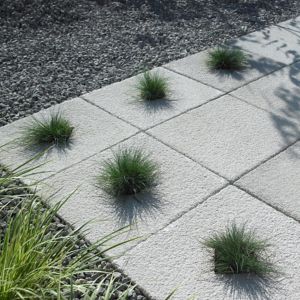 Textured grey paving slabs measuring 450x450x32mm, featuring a rough surface and arranged with small patches of grass between them, set against a gravel background.