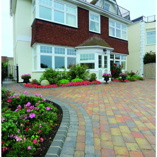 Bradstone Woburn rumbled concrete block paving in autumn hues, featuring a mix of warm reds, browns, and greys, arranged in a patterned layout, surrounded by vibrant flower beds.