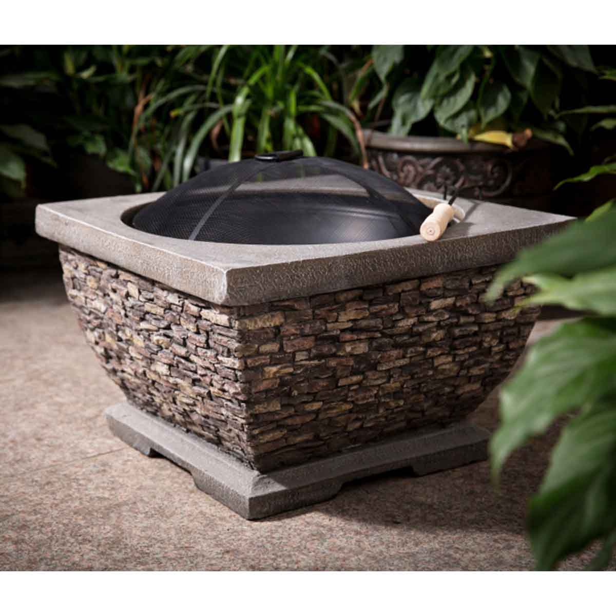 Callow Premium Wood Burner Fire Pit features a stone-textured base in gray with a round black metal fire bowl and a mesh spark guard, surrounded by lush green plants.