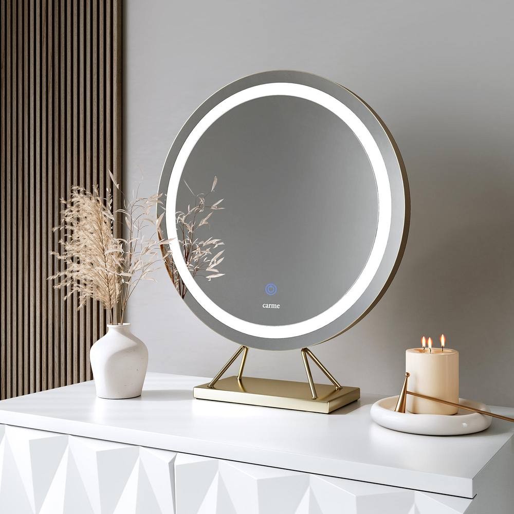 Frameless round gold mirror with touch sensor LED light, set on a sleek gold stand, displayed on a white surface alongside a vase of dried flowers and a lit candle.
