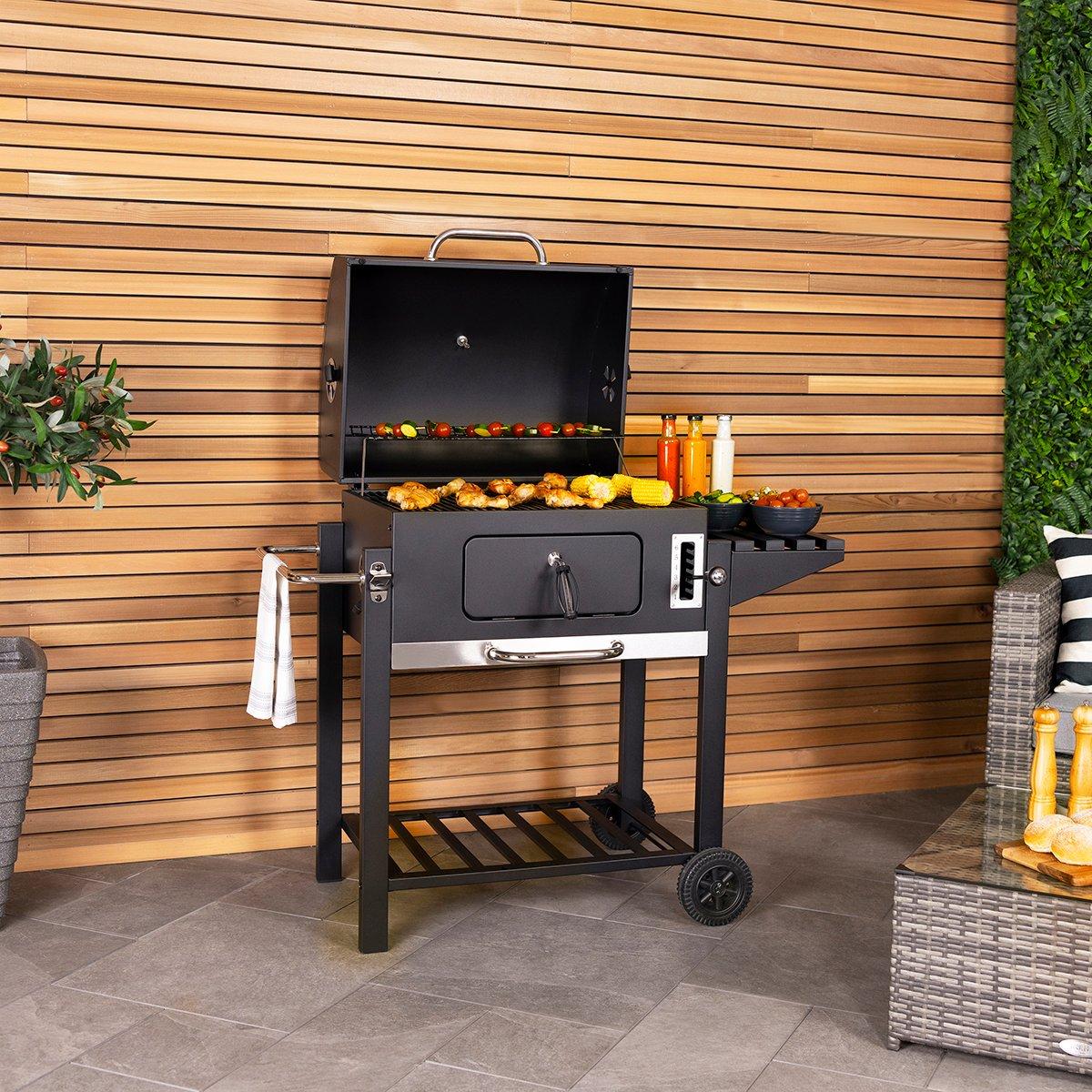 Large black charcoal BBQ grill with a 60x45cm cooking area, featuring a spacious lid, side shelves, and wheels for portability, set against a wooden backdrop with greenery.