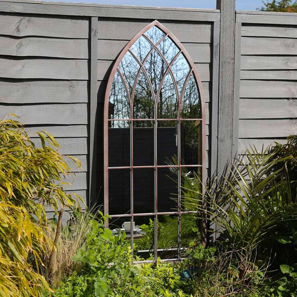 Grey arch-shaped outdoor mirror with a decorative lattice design, framed in grey metal, set against a wooden fence and surrounded by greenery.