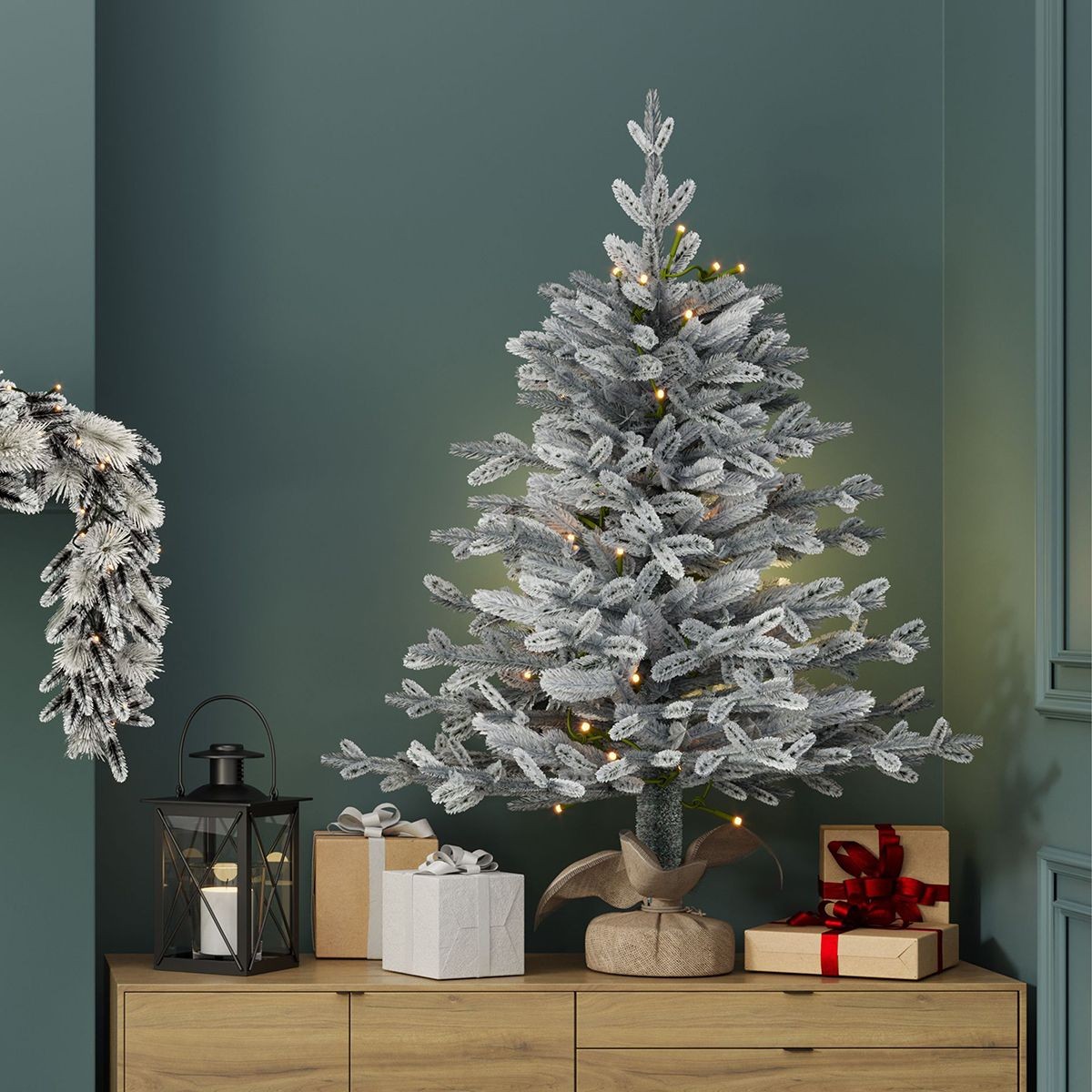 Luxury 3ft pre-lit faux snowy Christmas tree with frosted branches, warm white lights, and a burlap base, displayed beside wrapped gifts and a lantern against a green wall.
