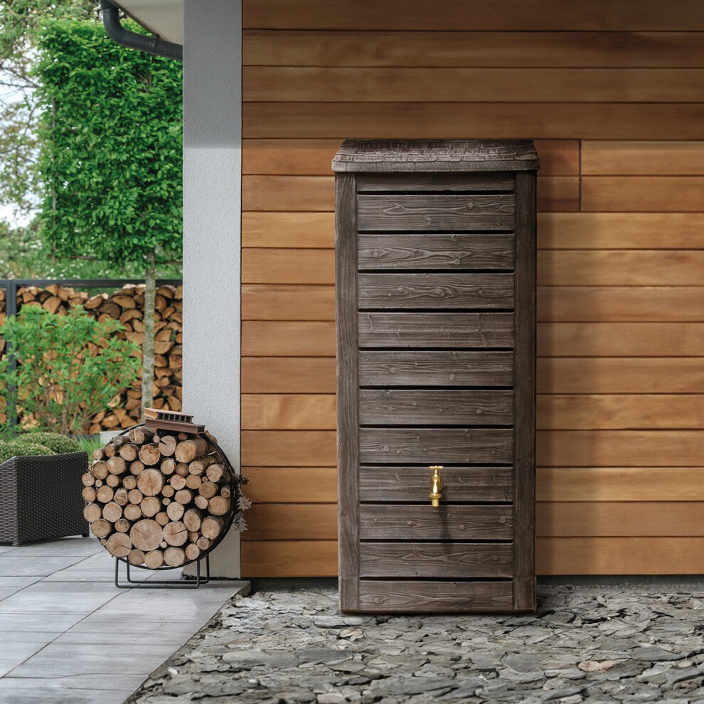 Brown wooden water butt with a textured finish, featuring a brass tap at the bottom, positioned against a wooden wall with stacked logs nearby.