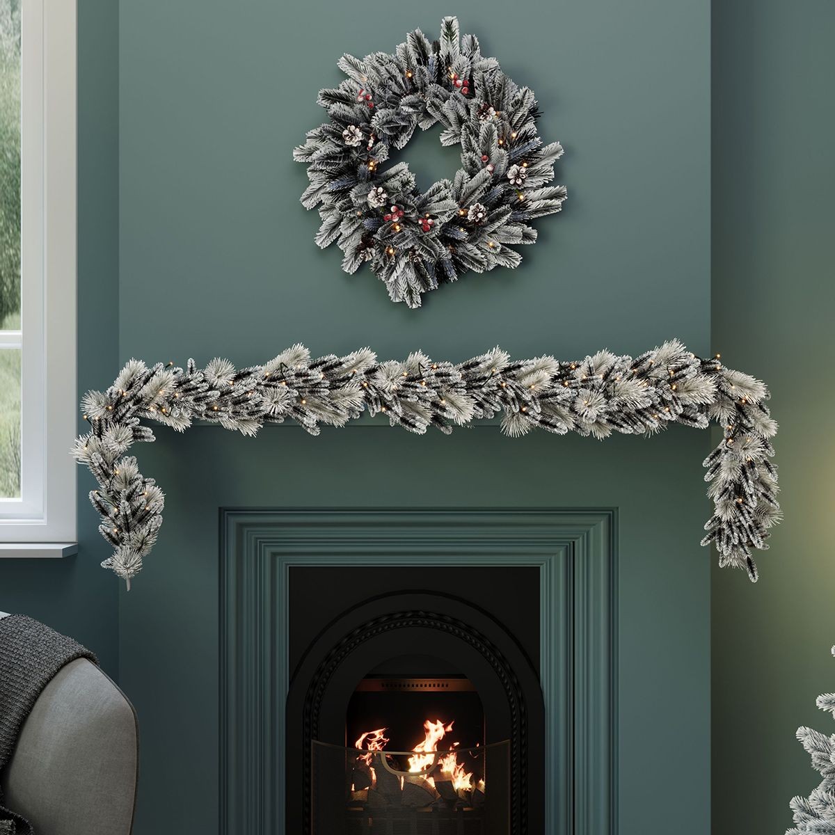 Snowy green pine garland adorned with red berries and pinecones, featuring warm white lights, draped elegantly above a fireplace, complemented by a matching wreath above.