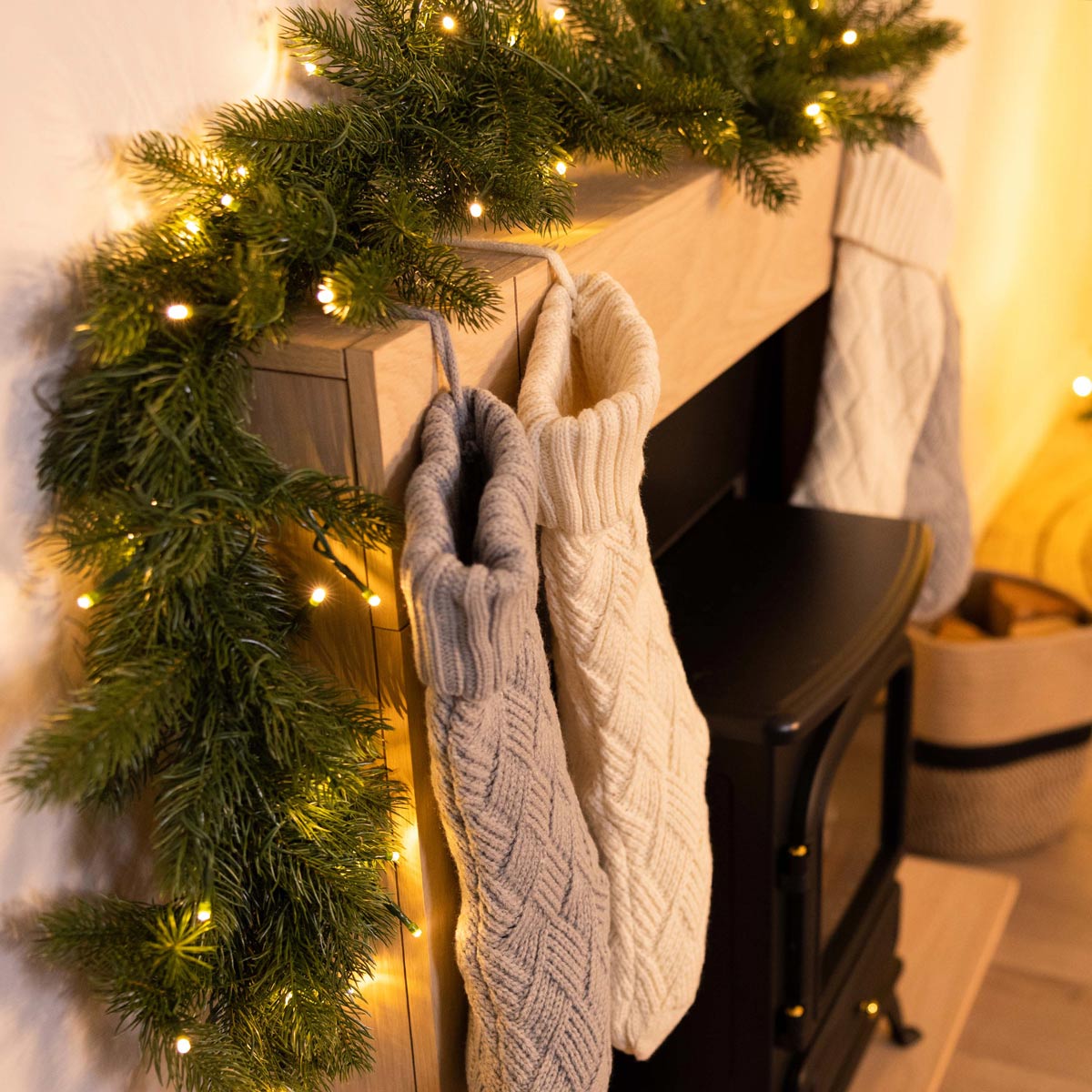 Faux green Christmas garland, 180cm long, adorned with warm white lights, draped over a wooden mantel, with knitted stockings hanging below.