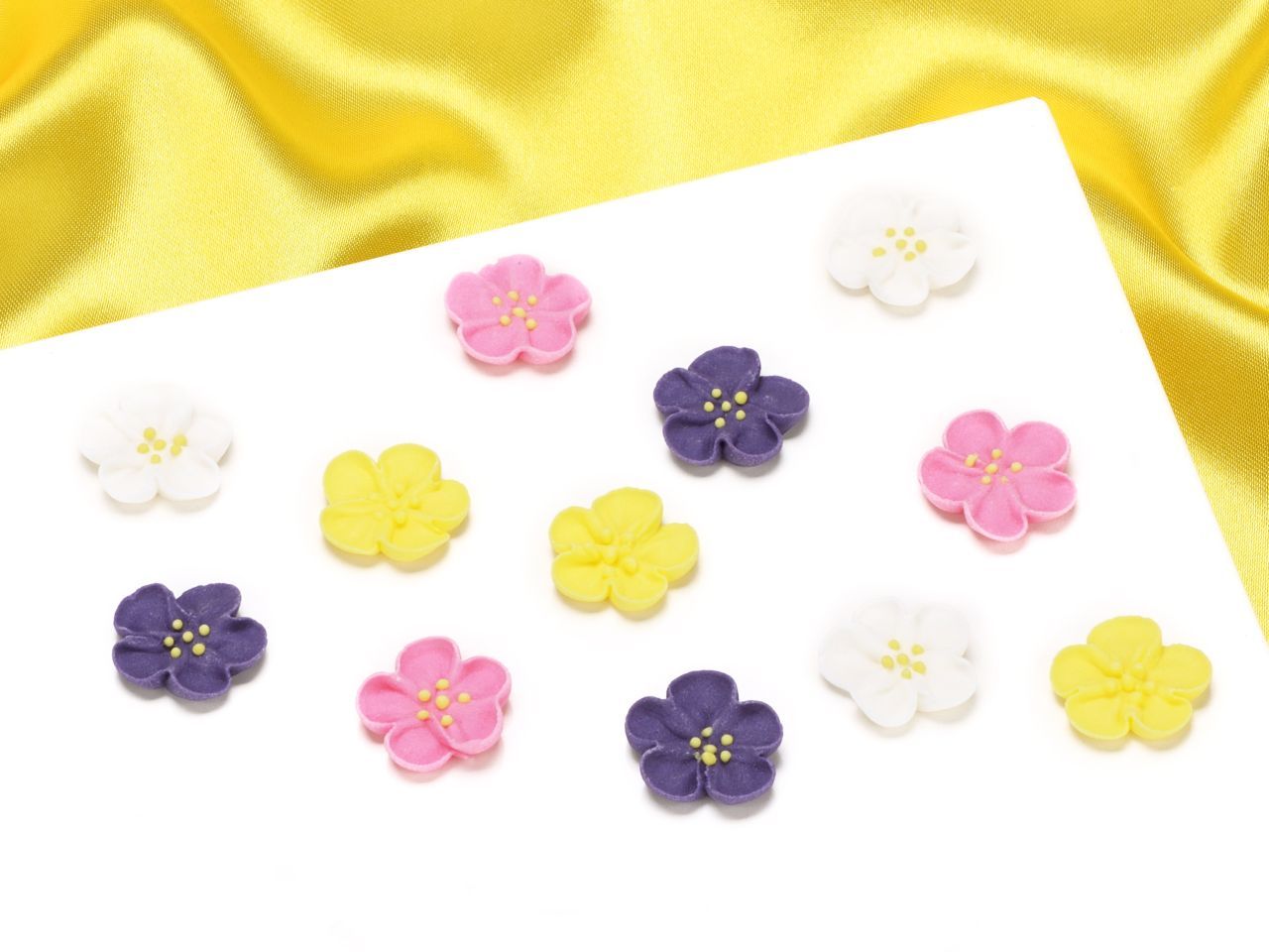 Colorful edible sugar decorations featuring 12 small flowers in pink, purple, yellow, and white, each with detailed petals and tiny sugar bead centers, set against a satin background.