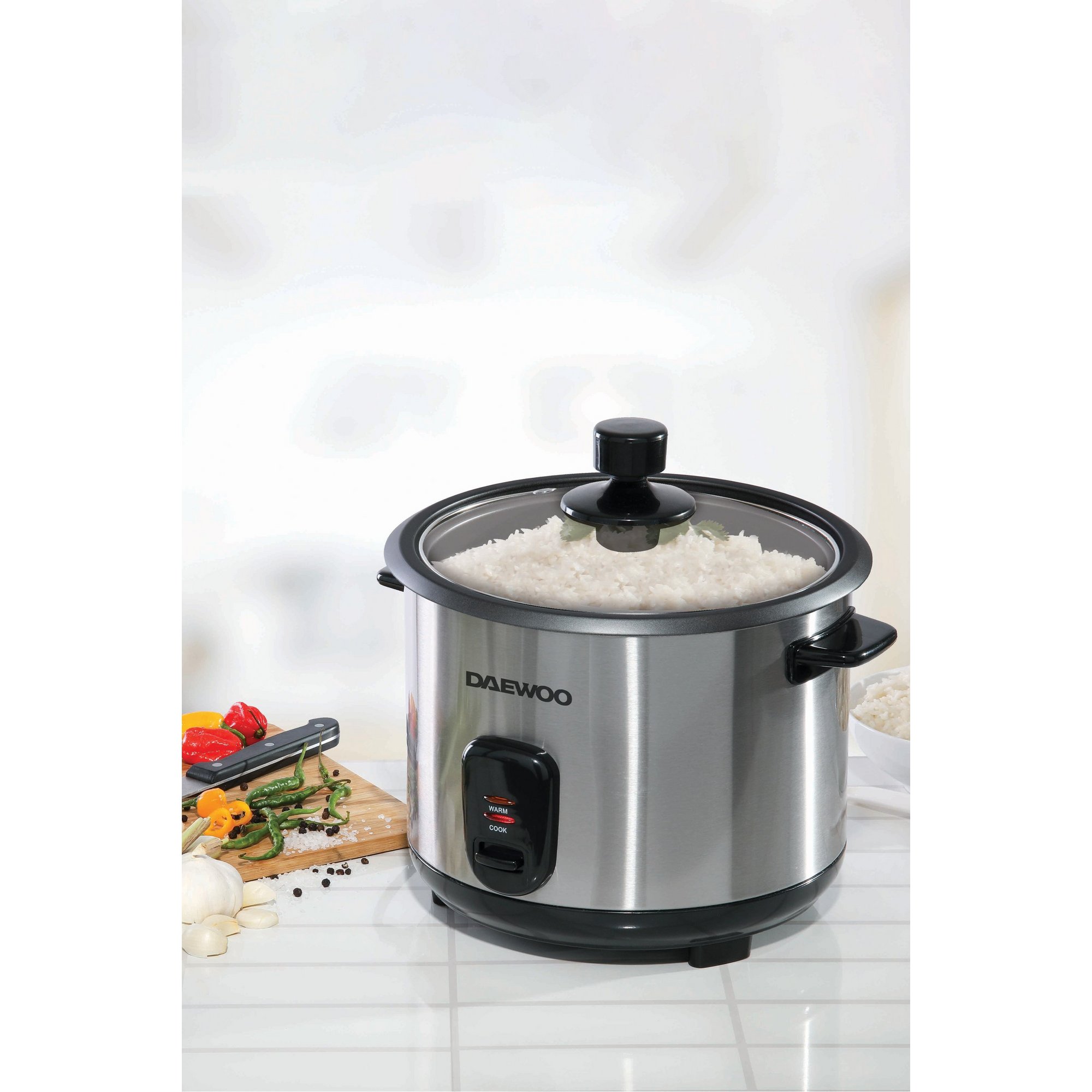 Black Daewoo rice cooker with a 1.8-litre capacity, featuring a stainless steel body, transparent lid, and a red indicator light, surrounded by fresh vegetables and spices.