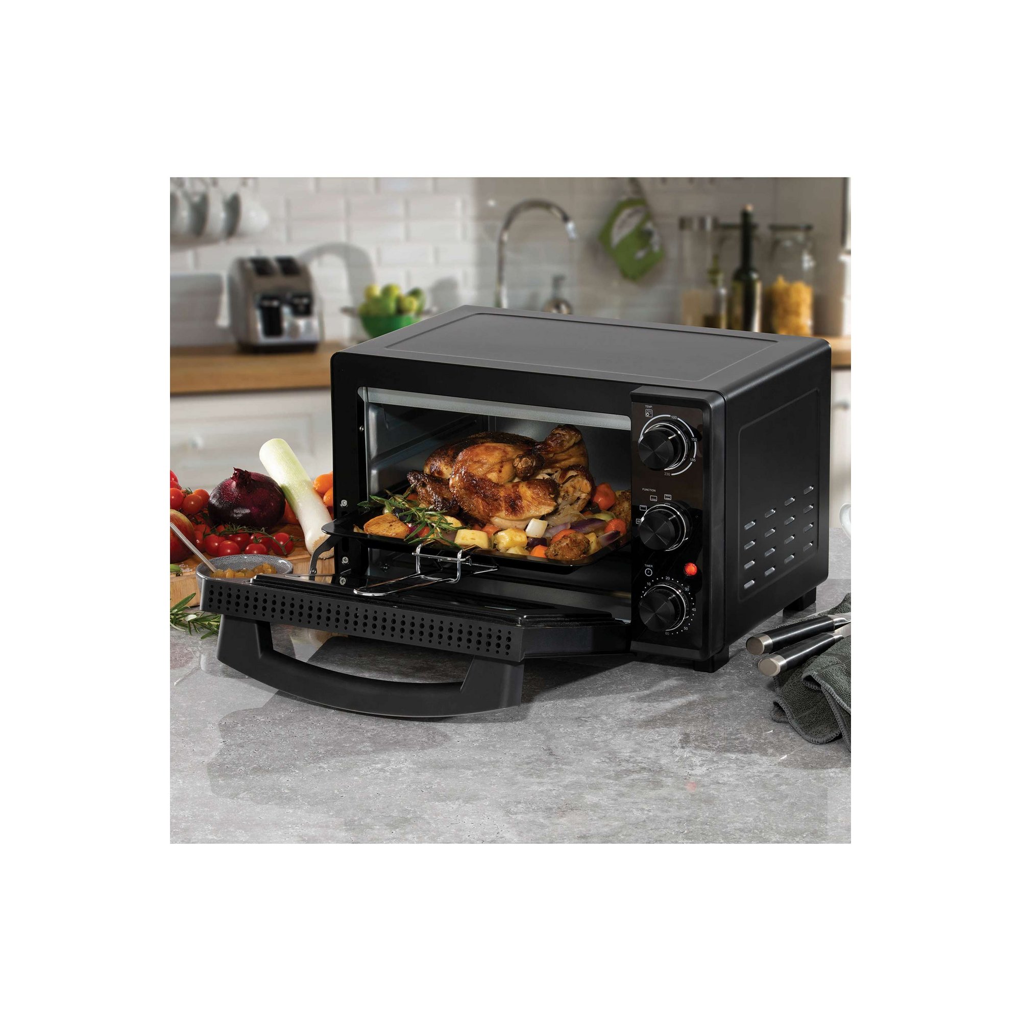 Black Daewoo 23L electric mini oven with adjustable temperature control (90°C - 230°C), timer dial, and a pull-out tray, set on a kitchen countertop surrounded by fresh vegetables.