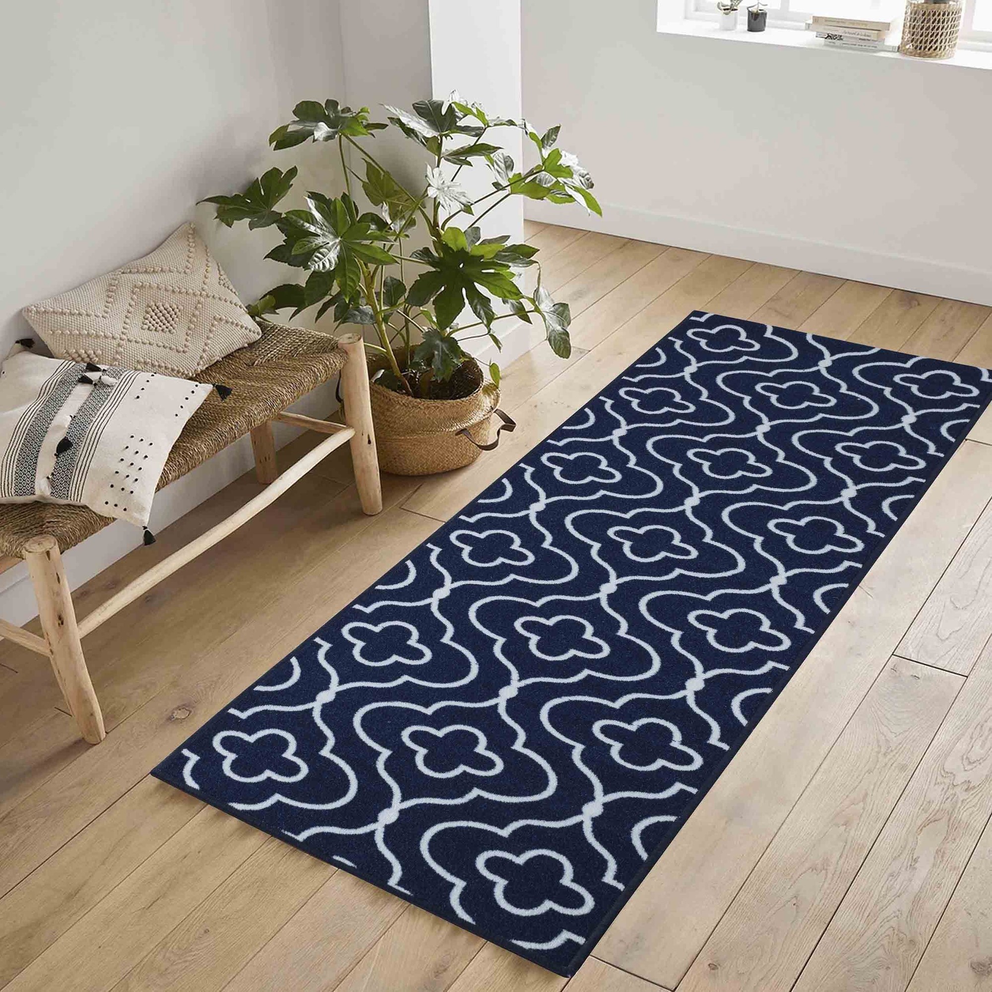 Navy blue doormat featuring a white anti-slip quatrefoil design, made from machine-washable material, placed on a wooden floor beside a potted plant and a chair.