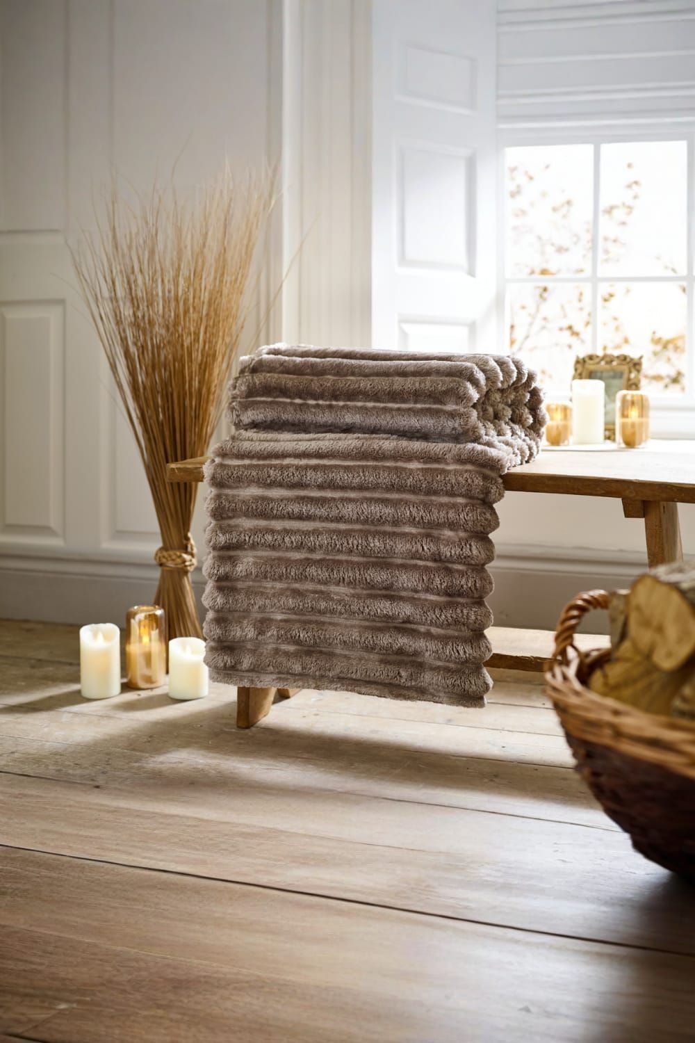 Taupe ribbed throw blanket stacked on a wooden table, featuring a soft plush texture, with candles and decorative plants in a cozy room setting.