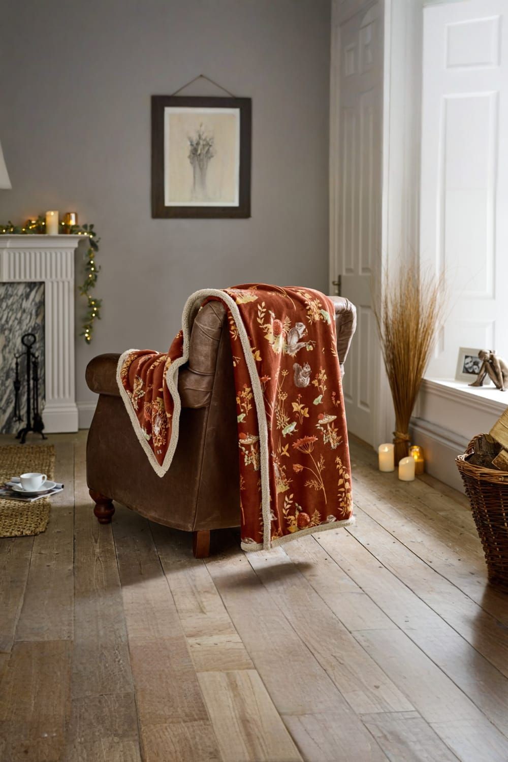 Rust-colored Sherpa-lined blanket featuring woodland animals and floral patterns, draped over a brown armchair in a cozy, warmly lit room with candles and decor.
