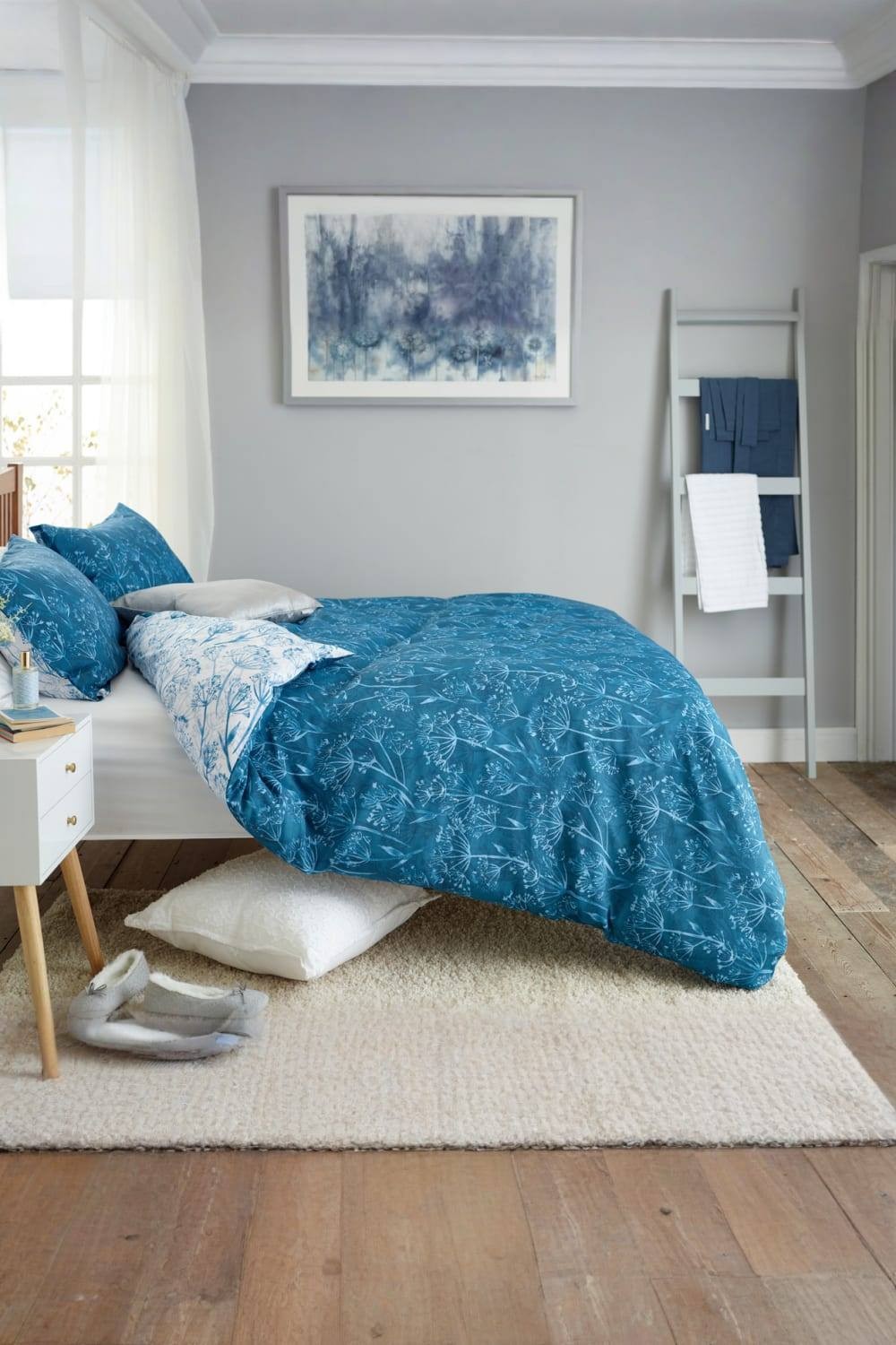 Blue cotton-rich duvet cover set featuring a cow parsley print, with matching pillowcases, displayed on a single bed in a softly lit room with wooden flooring.