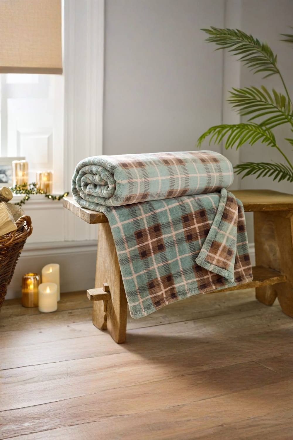 Ultra-soft green and brown Highland Fell check throw blanket, measuring 140x180cm, neatly rolled on a wooden bench with candles and a plant in the background.