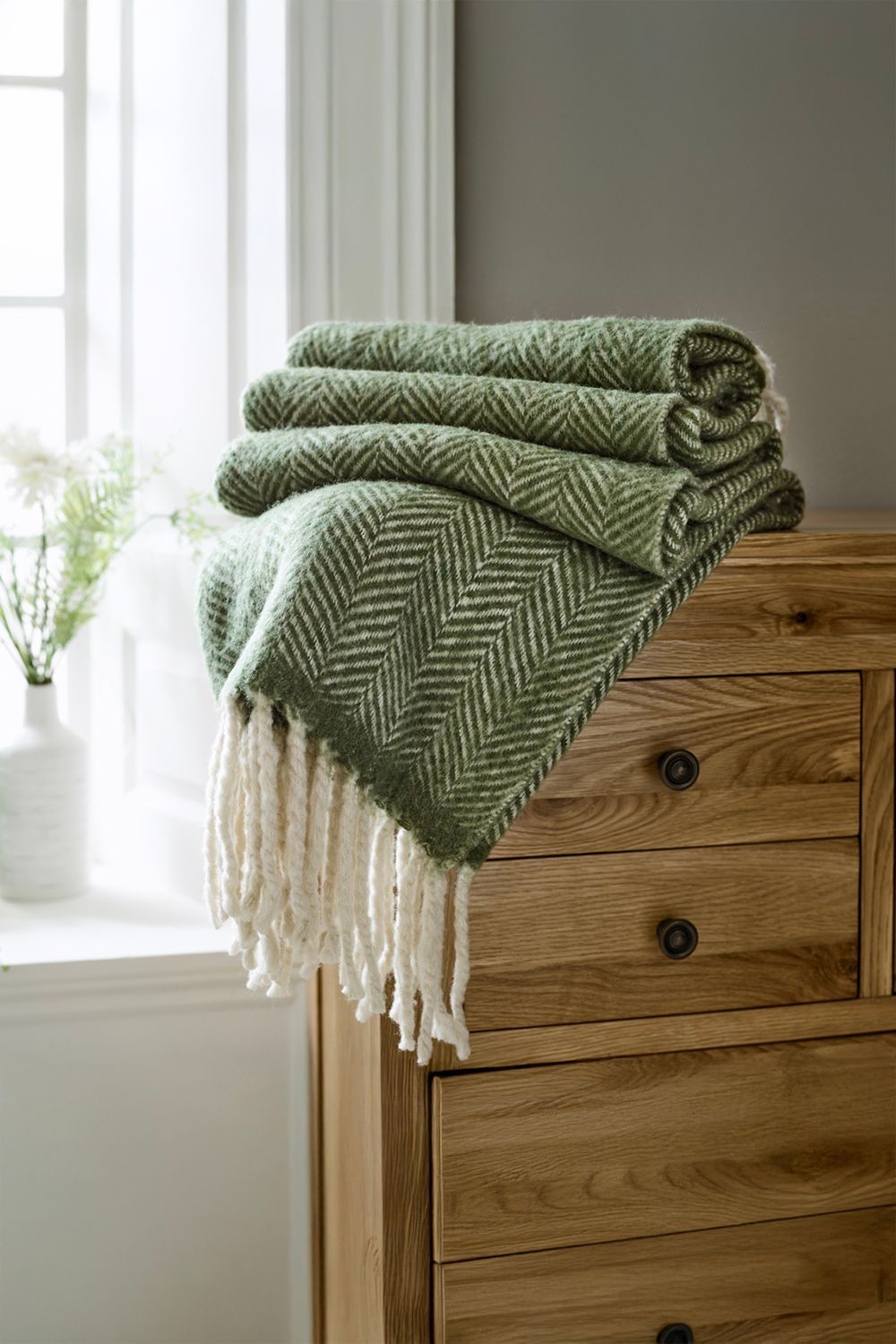 Supersoft green jacquard throw made of 100% brushed wool, featuring a herringbone pattern and cream fringes, neatly folded on a wooden dresser near a window.