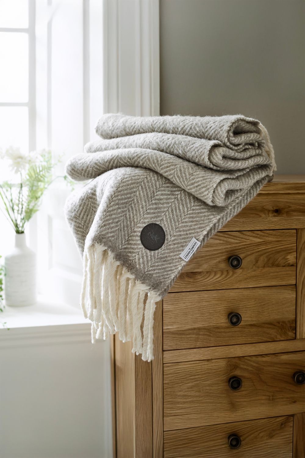 Supersoft natural-colored jacquard throw made from 100% brushed wool, featuring a herringbone pattern and fringed edges, neatly folded on a wooden dresser.