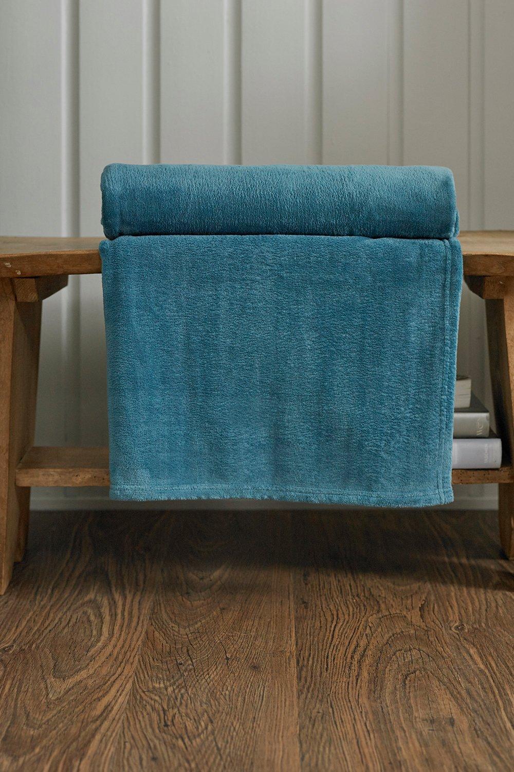 Soft, teal "Snuggle Touch" blanket by Deyongs, featuring a plush texture, neatly rolled at the top, draped over a wooden bench with a rustic finish.