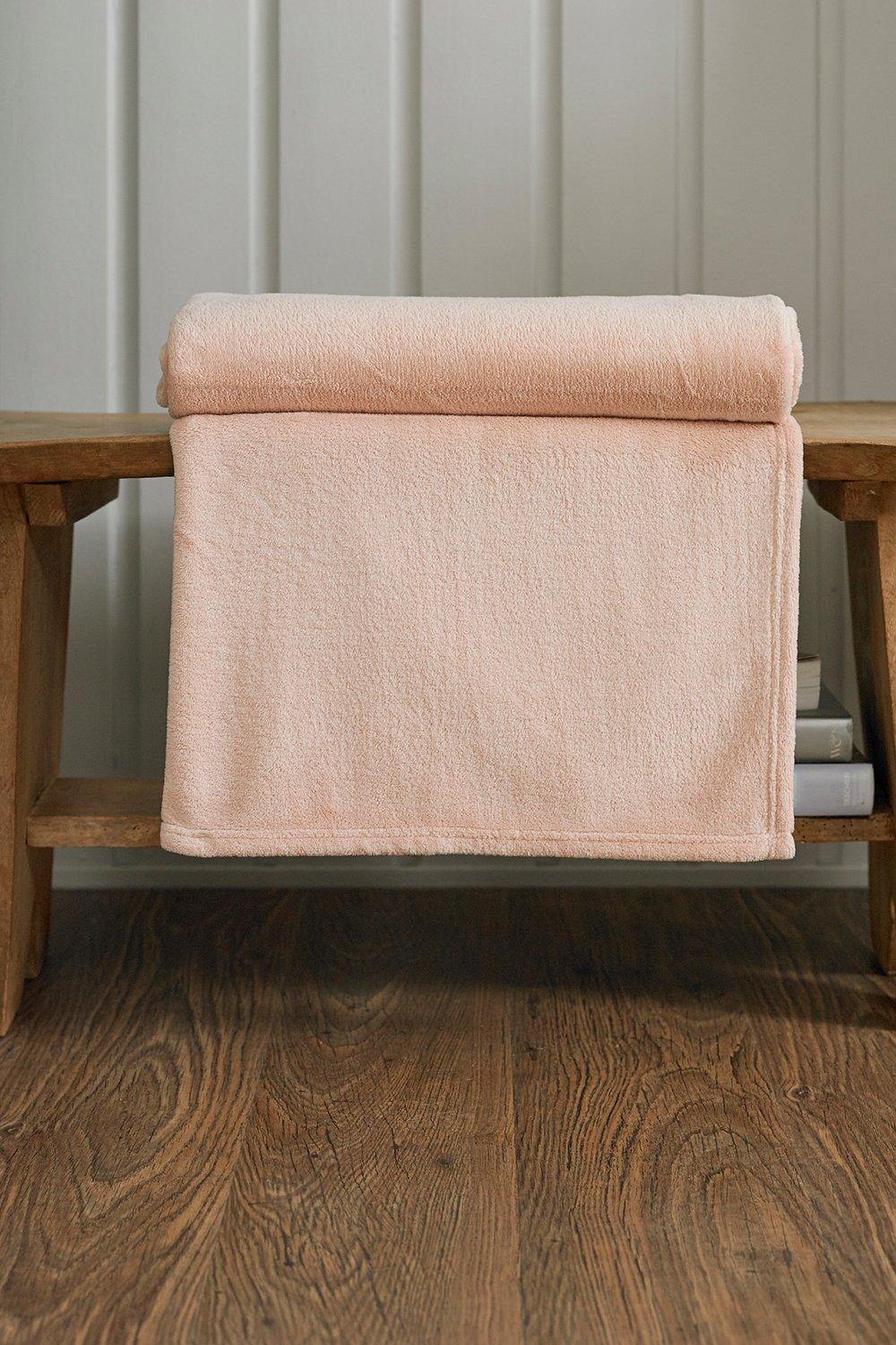 Soft pink microfibre throw measuring 140x180cm, neatly rolled and draped over a wooden bench, showcasing its plush texture and cozy appearance.