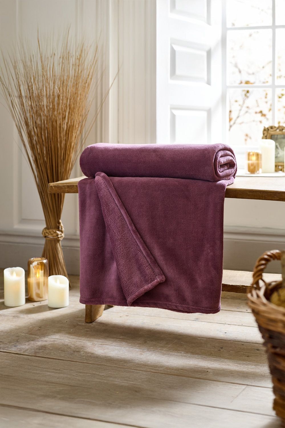 Magenta fleece throw by Deyongs, measuring 140cm x 180cm, displayed neatly on a wooden bench with candles and decorative elements in a cozy setting.