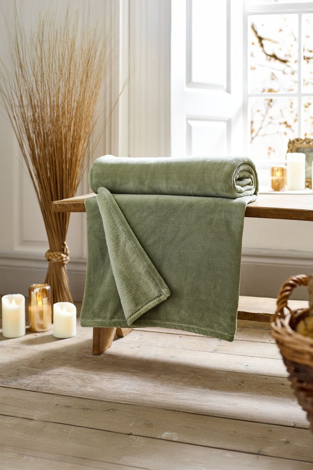Soft green fleece throw rolled and draped on a wooden bench, with candles and decorative plants in the background, showcasing its cozy texture and solid color.