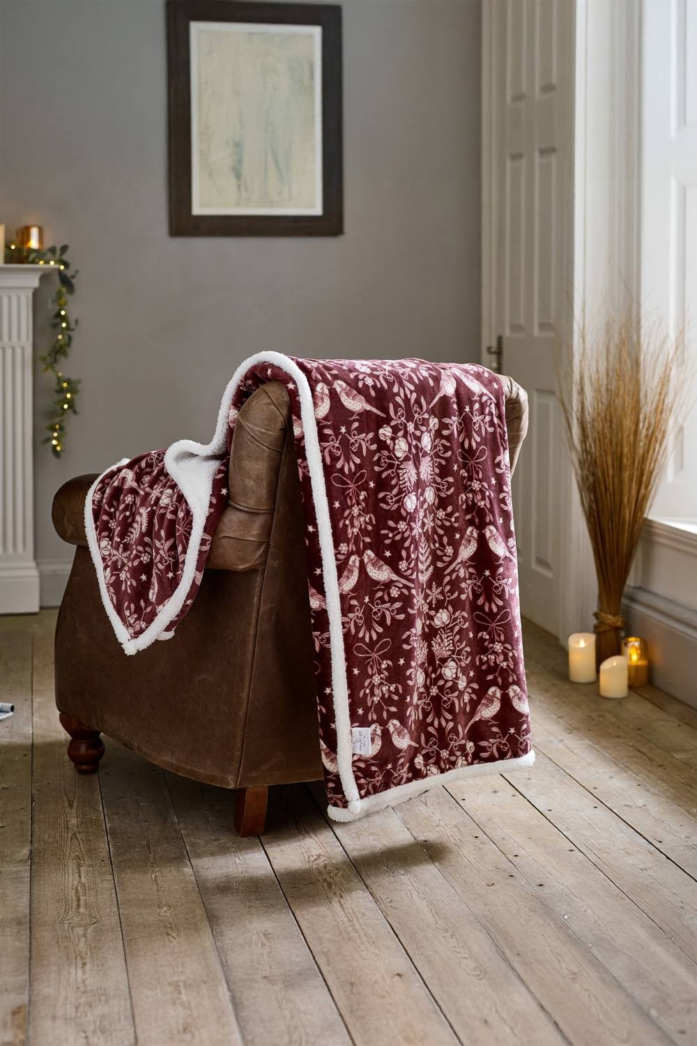 Burgundy fleece throw with a bird and floral pattern, featuring a soft Sherpa reverse, draped over a brown leather armchair in a cozy indoor setting.