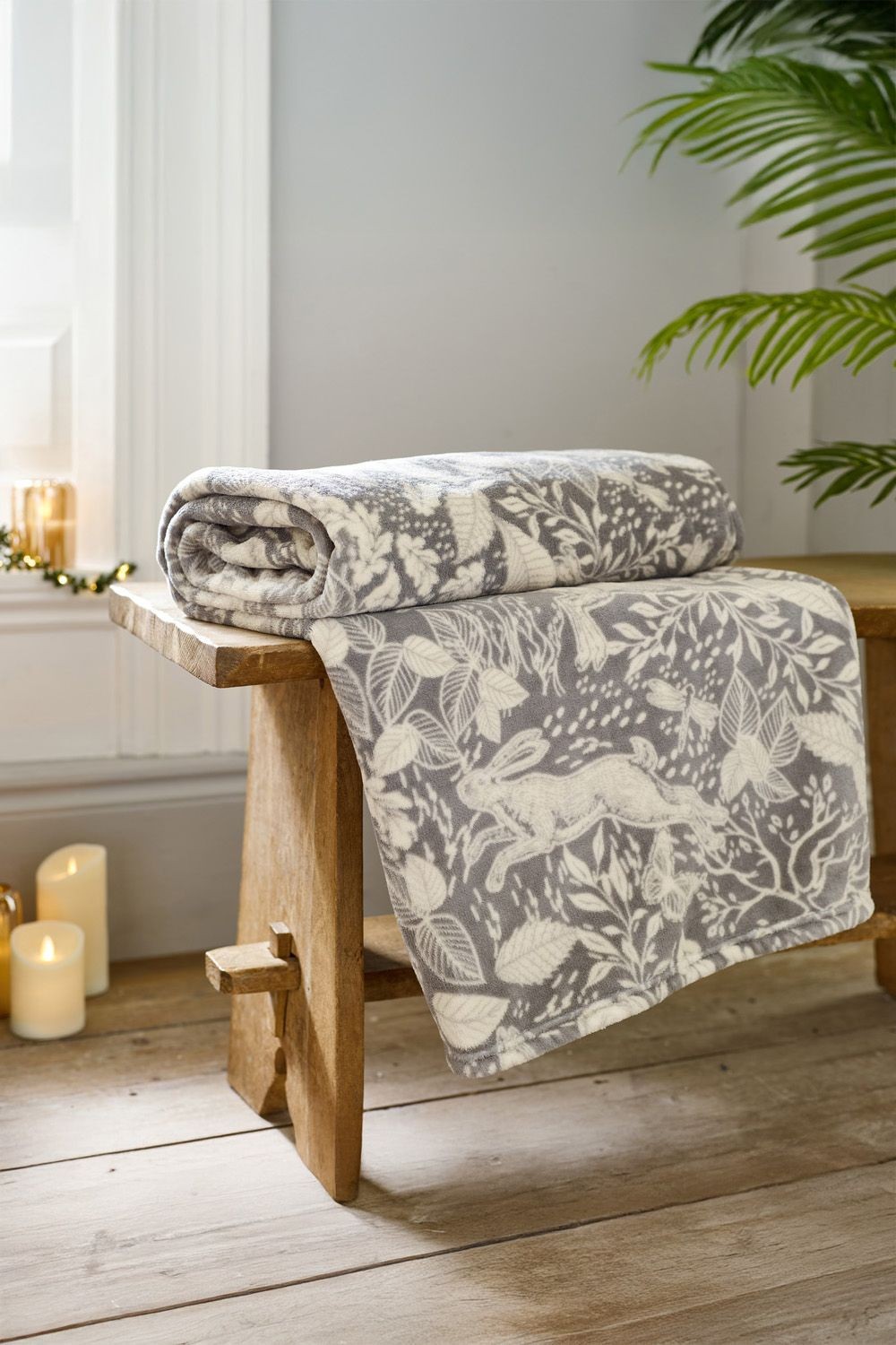 Soft grey fleece throw blanket featuring a woodland animal design, rolled neatly on a wooden bench, with decorative candles and a plant in the background. Size: 140x180cm.