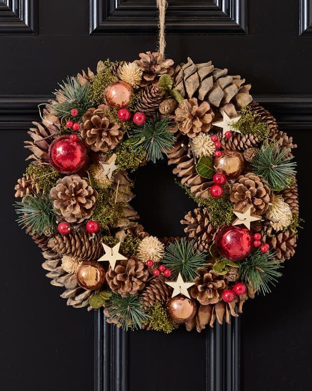 A 35cm Christmas wreath featuring natural pinecones, green foliage, red baubles, and wooden stars, accented with festive red berries and a rustic twine hanger.