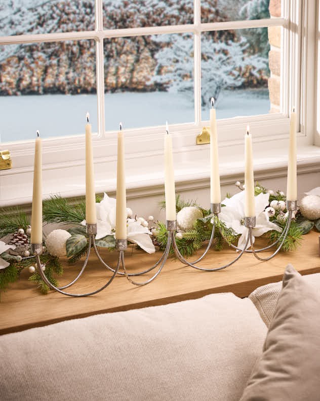 Elegant silver wave taper candle holder with six slender white candles, set against a wintery window view, adorned with greenery and decorative accents.