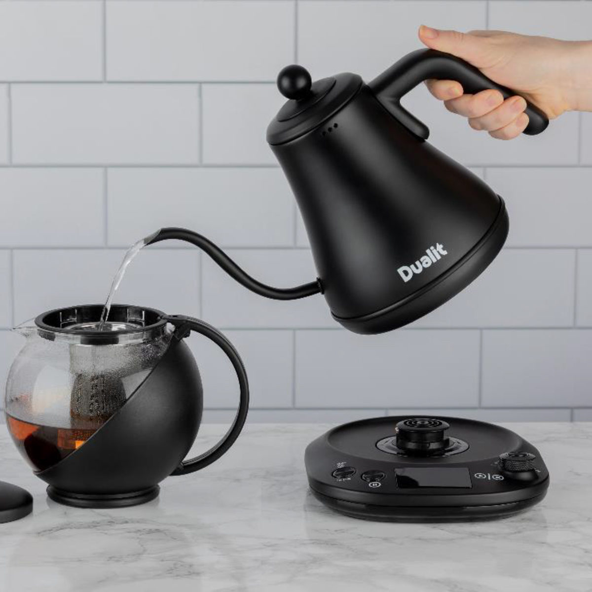 Black Dualit 0.8L pour-over kettle with a precision spout, being used to pour water into a glass teapot on a sleek, modern scale.