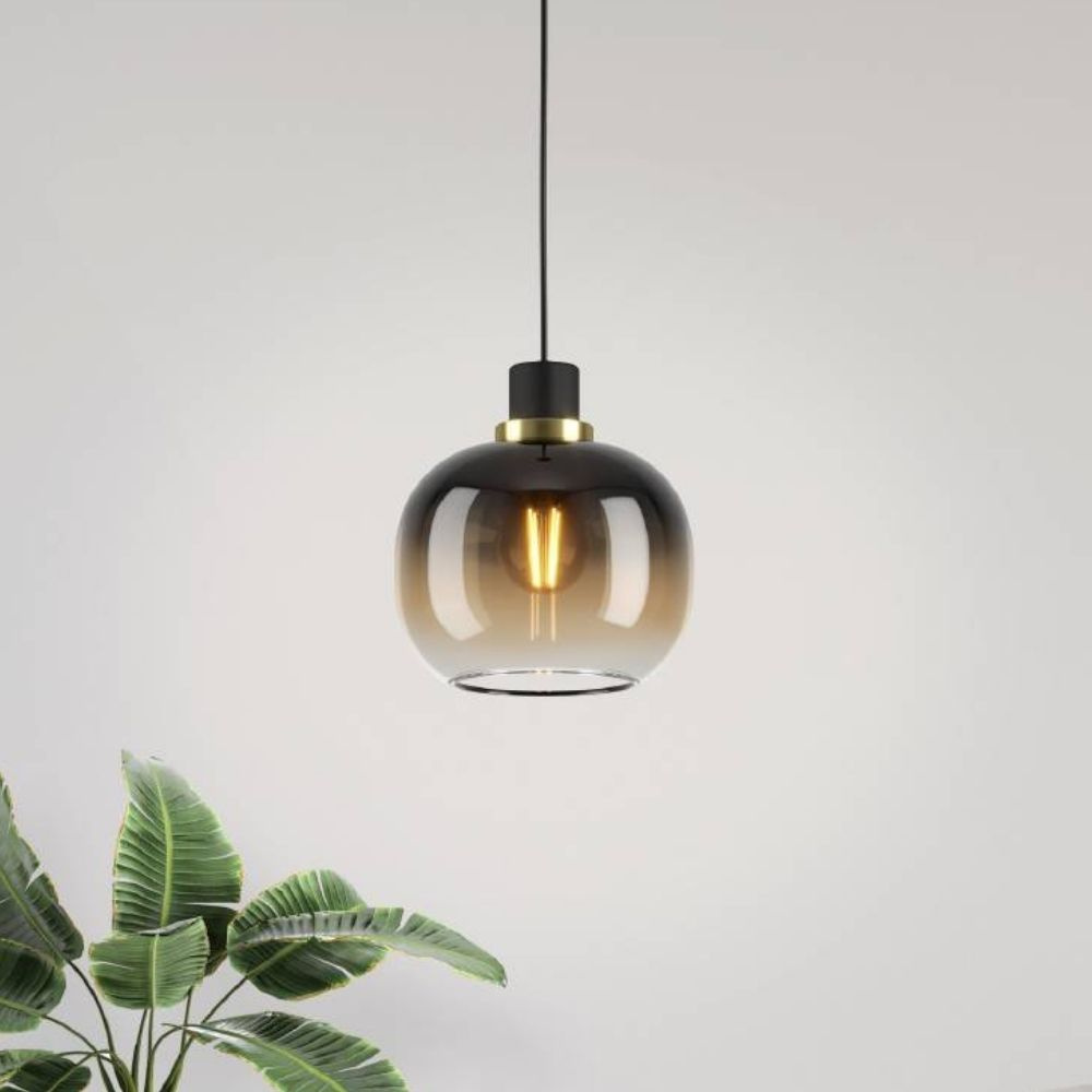 Light brown glass pendant light with a rounded shape, featuring a black metal top and an exposed bulb, suspended from a black cord, complemented by green foliage.