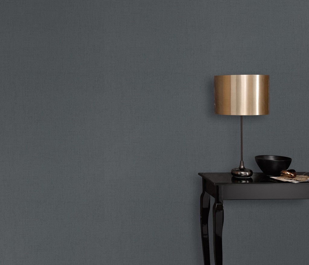 Textured dark grey wallpaper by Erismann, featuring a smooth finish, paired with a sleek black table, a gold lamp, and a black bowl on top.