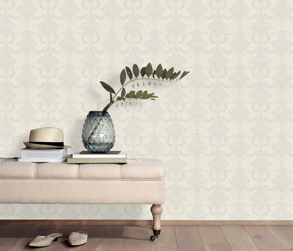 Light beige Vliestapete with a subtle damask pattern, displayed in a stylish interior featuring a tufted ottoman, a glass vase with greenery, and stacked books.