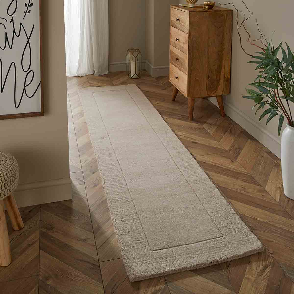 Cream wool runner measuring 60x230cm, featuring a simple design with a subtle border, placed on a herringbone wooden floor beside a wooden dresser and decorative elements.
