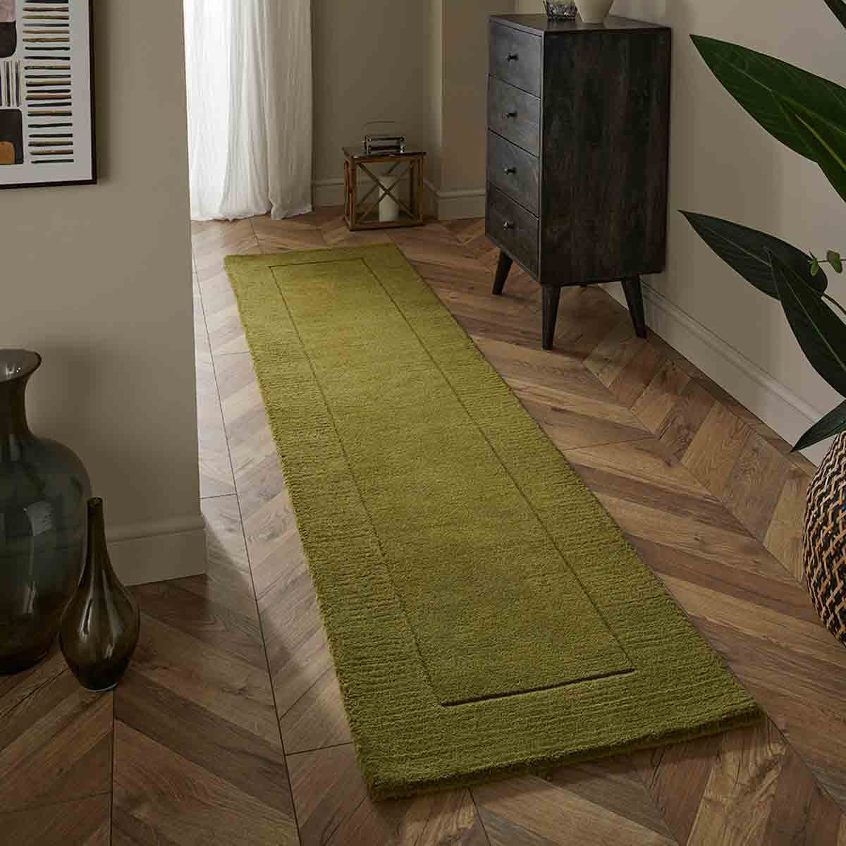 Olive green wool runner measuring 60x230cm, featuring a textured surface and bordered design, placed on a wooden floor beside a dark chest of drawers and decorative plants.