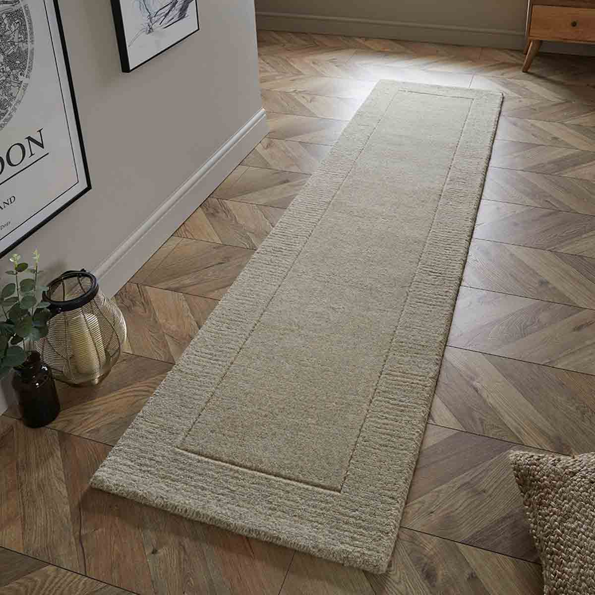 Mayfair Esme 60x230cm stone wool runner in a soft pink color, featuring a textured surface and bordered design, placed on a wooden floor in a well-lit room.
