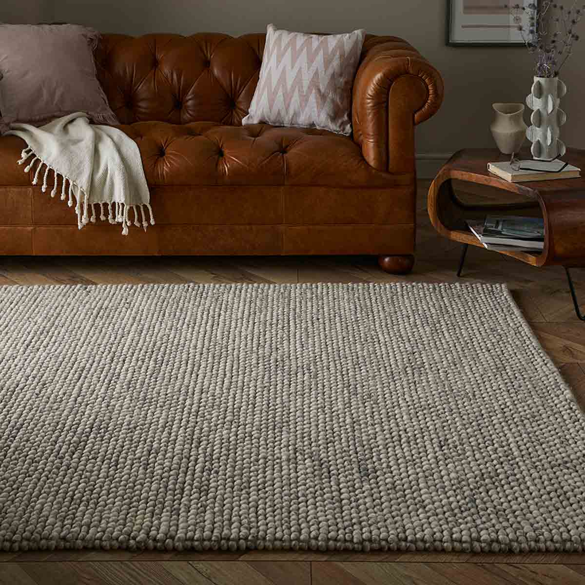 Mottled cream wool pebble rug measuring 160x230cm, featuring a textured surface, placed on a wooden floor near a brown leather couch and decorative pillows.