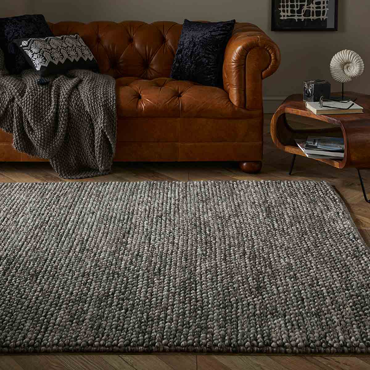 Mottled grey wool pebble rug measuring 160x230cm, featuring a textured surface, placed on a wooden floor in a cozy living room setting with a brown leather sofa and decorative cushions.