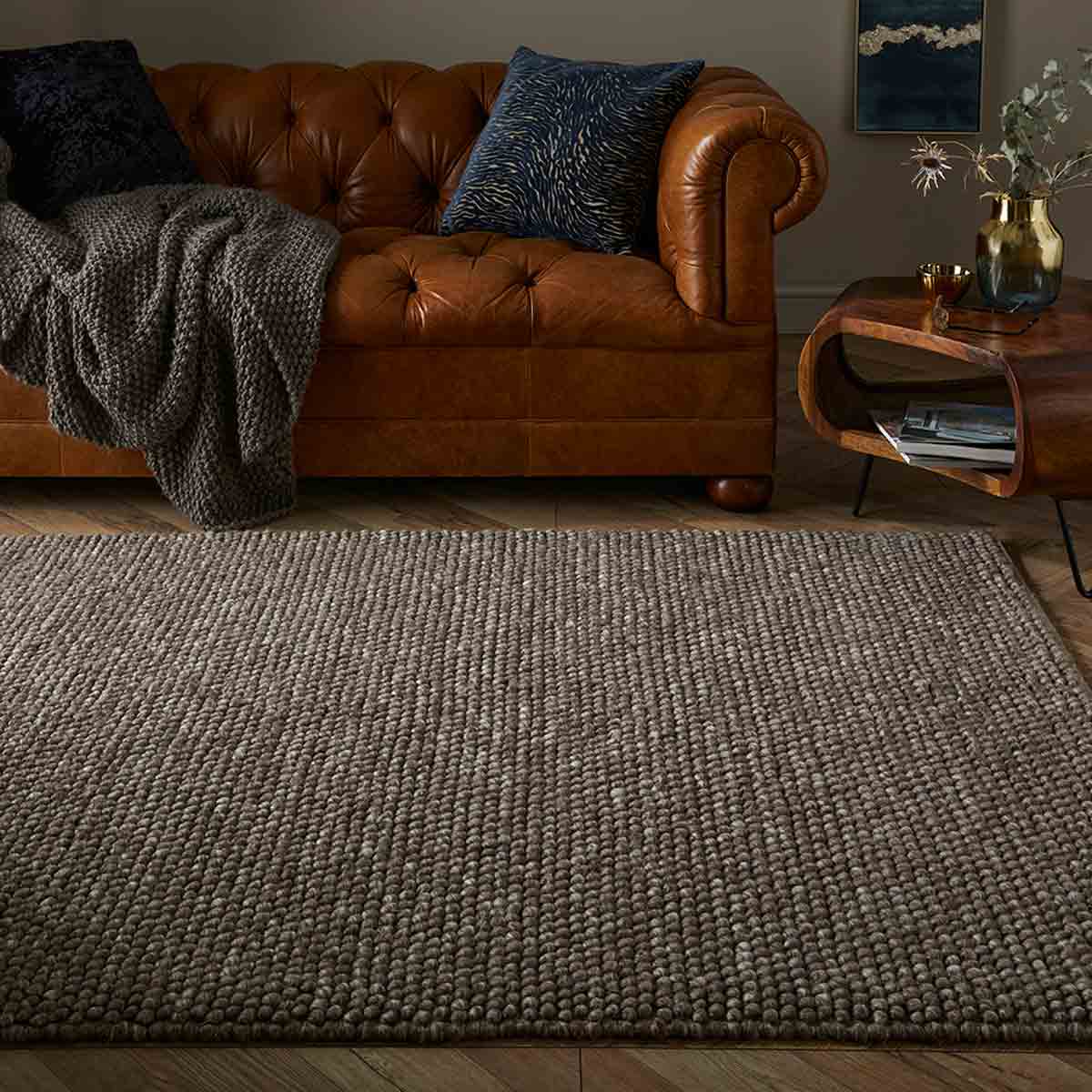 Mottled taupe wool pebble rug measuring 160x230cm, featuring a textured surface, placed on a wooden floor beside a brown leather sofa with decorative pillows.